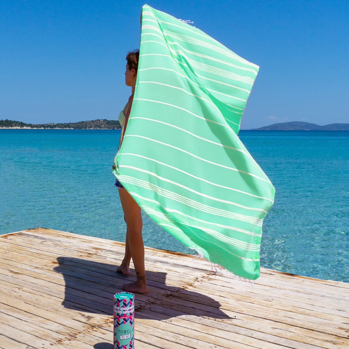 Frau hält grünes Towel To Go Hamamtuch im Wind auf einem Steg am Meer